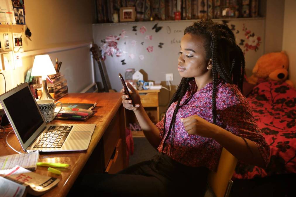 A young Black woman sits in her university dorm room looking at her phone with her laptop open on her desk.